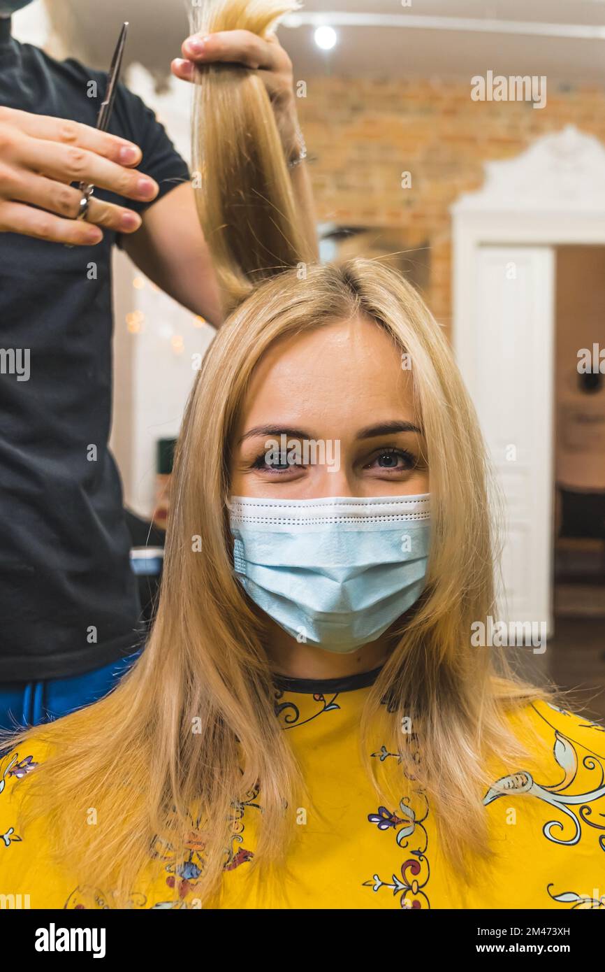 Donna bionda positiva in maschera protettiva che guarda la fotocamera durante un taglio di capelli. Verticale interno. Spazio di copia. Visita sicura del parrucchiere durante la pandemia. Foto di alta qualità Foto Stock