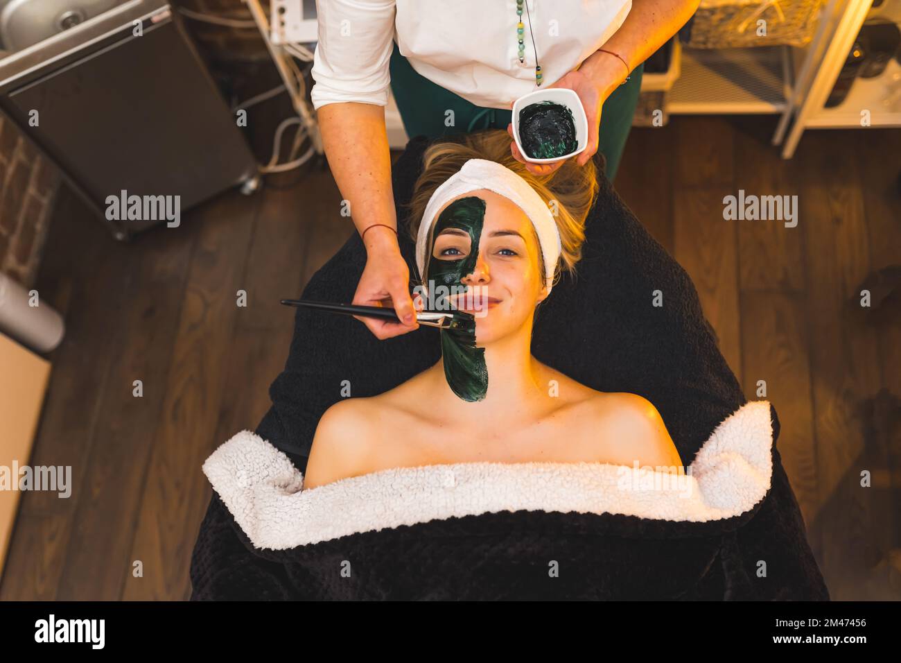 Cliente Happy SPA su letto SPA sotto una coperta con maschera al carbone. Estetista irriconoscibile che applica una maschera nera con un pennello. Scatto da interno ad angolo alto. Spazio di copia. Foto di alta qualità Foto Stock