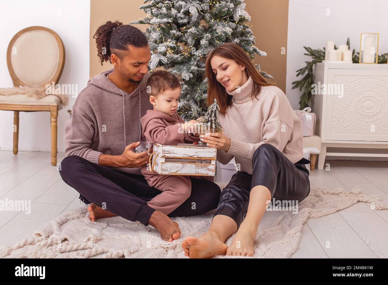 Una famiglia felice è seduta su una coperta a maglia, tenendo una scatola di legno con i giocattoli di Natale. Foto Stock