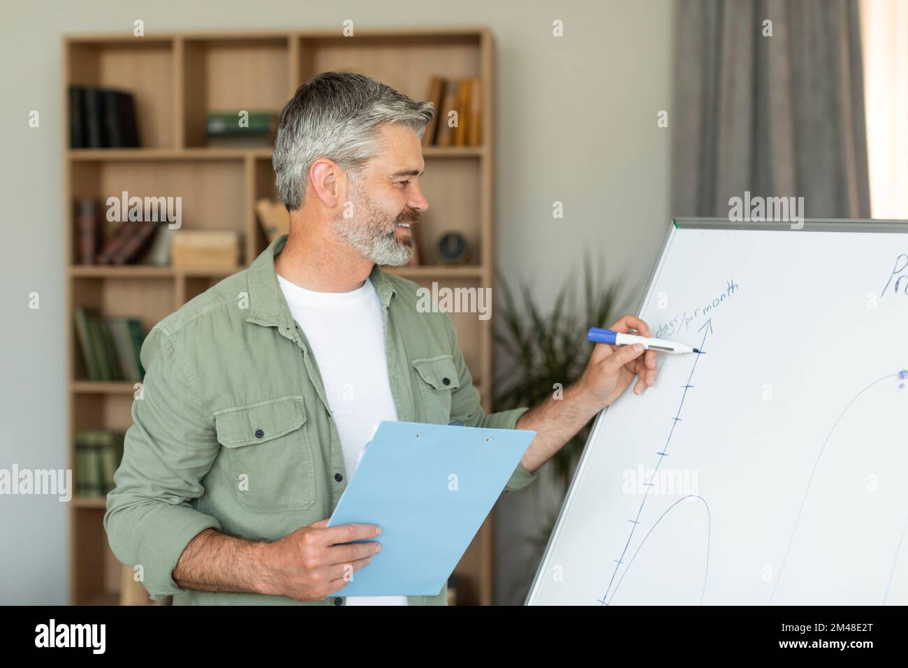 Sorridente anziano insegnante caucasico scrive sulla lavagna e registra la lezione video in salotto Foto Stock