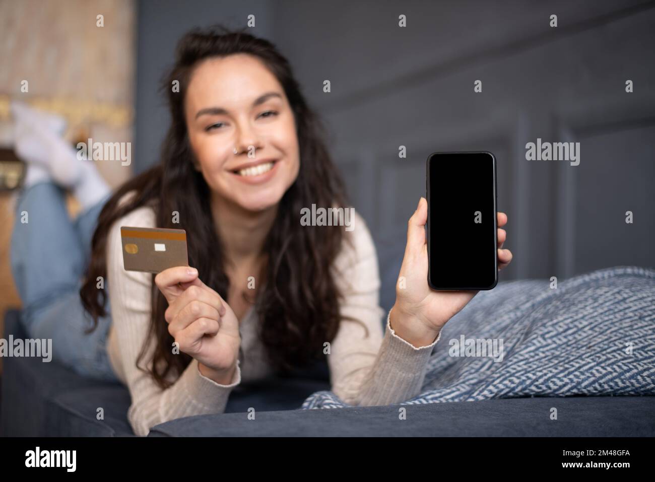 Donna caucasica felice mostra smartphone con schermo vuoto e carta di credito, sdraiato sul divano in soggiorno, mockup Foto Stock