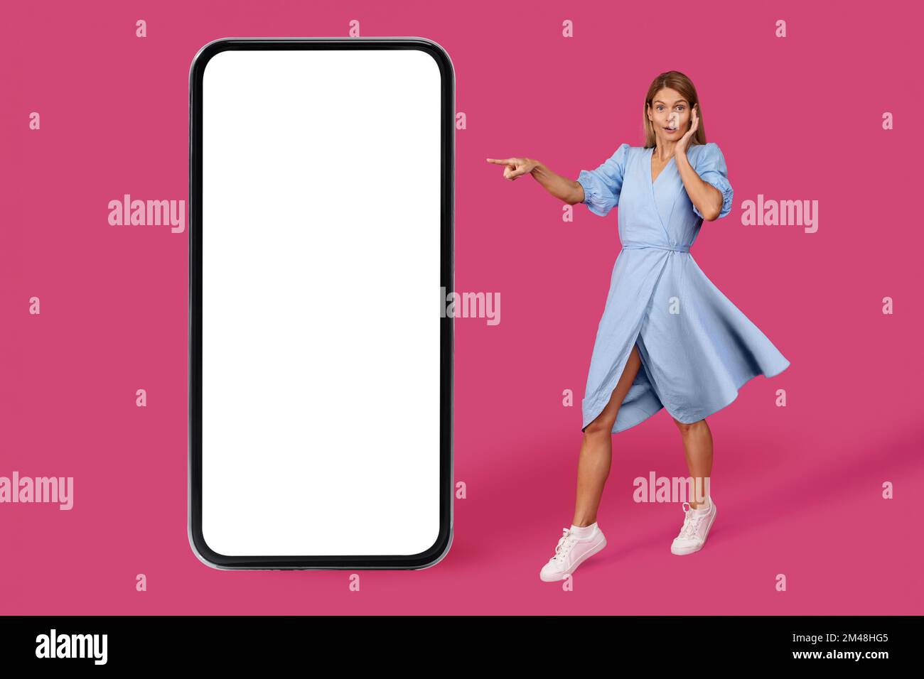 Adulta scioccata donna caucasica bionda in vestito punto dito a grande smartphone con schermo vuoto Foto Stock