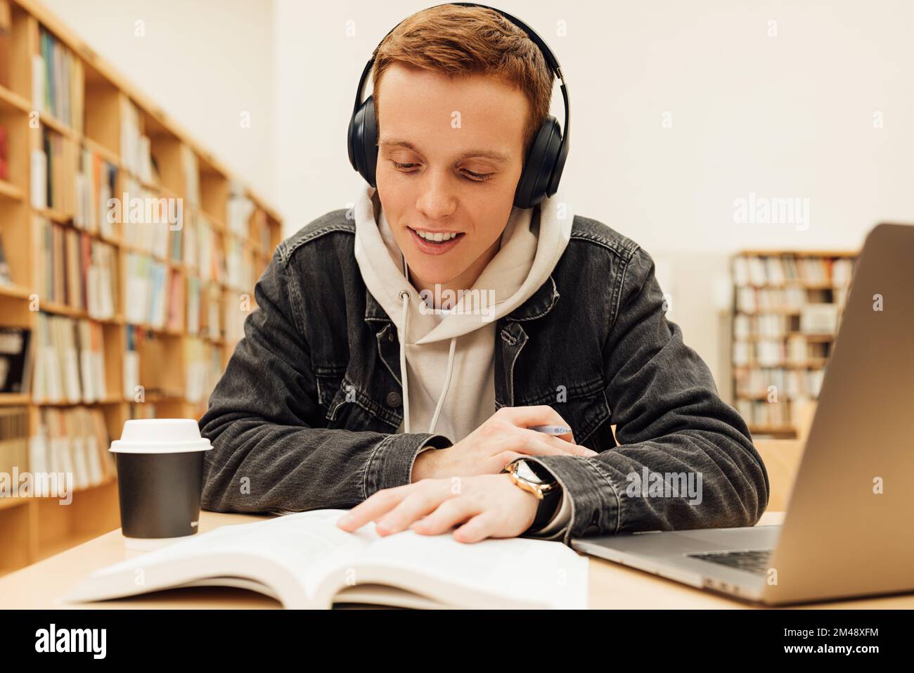 Sorridente studente che legge un libro indossando le cuffie mentre si siede in una biblioteca Foto Stock