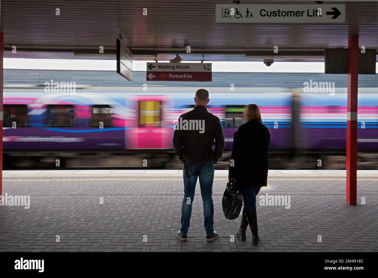 I passeggeri del treno che aspettano alla stazione ferroviaria di Wigan North Western sulla linea principale della costa occidentale con un treno che supera la velocità senza sosta Foto Stock