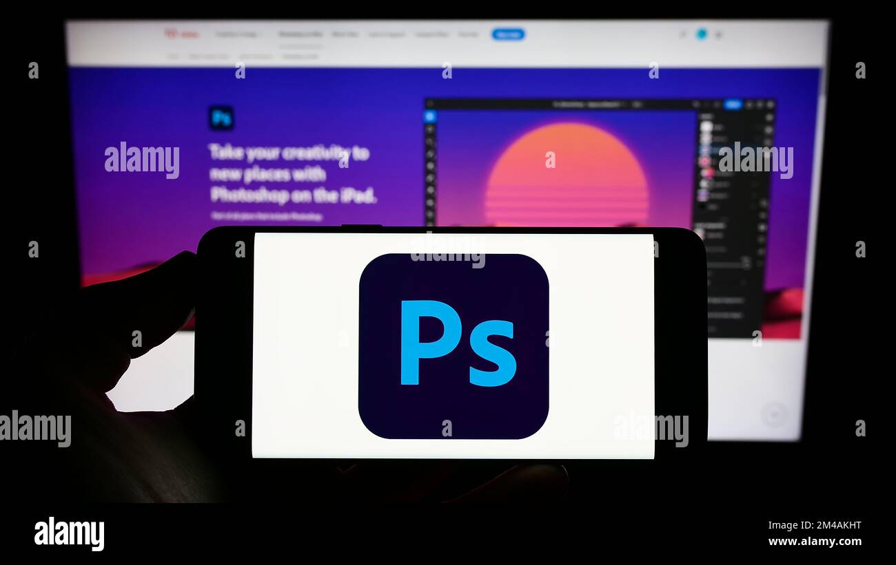 Persona che tiene il cellulare con il logo del software di editor grafico Adobe Photoshop sullo schermo di fronte al sito web aziendale. Messa a fuoco sul display del telefono. Foto Stock