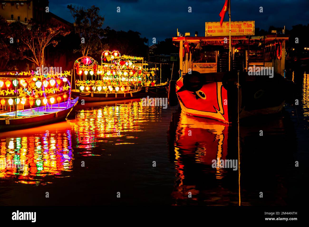 Hoi An tour delle barche della città vecchia di notte con lanterne colorate appese sulle barche che gettano riflessi accanto a uno scafo rosso barca e una linea ormeggio. Foto Stock