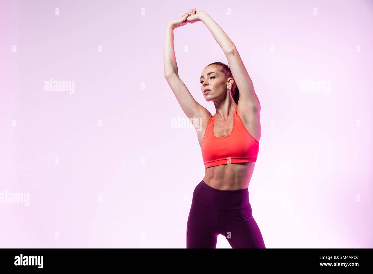 Sportivo adulto in abbigliamento sportivo che solleva le braccia e si piega da parte mentre si scalda durante l'allenamento fitness su sfondo rosa Foto Stock