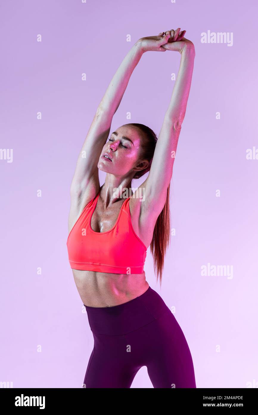 Sportivo adulto in abbigliamento sportivo che solleva le braccia e si piega da parte mentre si scalda durante l'allenamento fitness su sfondo lilla Foto Stock