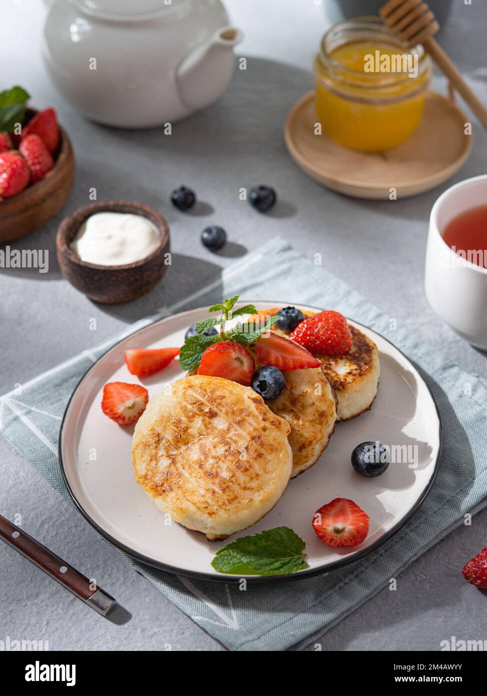 Frittelle con formaggio casolare o sciroppi con mirtilli freschi, fragole, miele e panna acida su sfondo blu con una tazza di tè e un bollitore Foto Stock