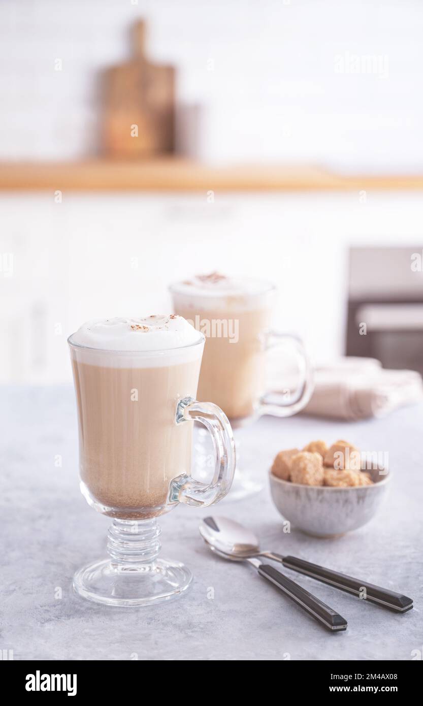 Due bicchieri di fragrante cappuccino con cannella e zucchero di canna su un tavolo blu sullo sfondo di una cucina bianca al mattino presto Foto Stock