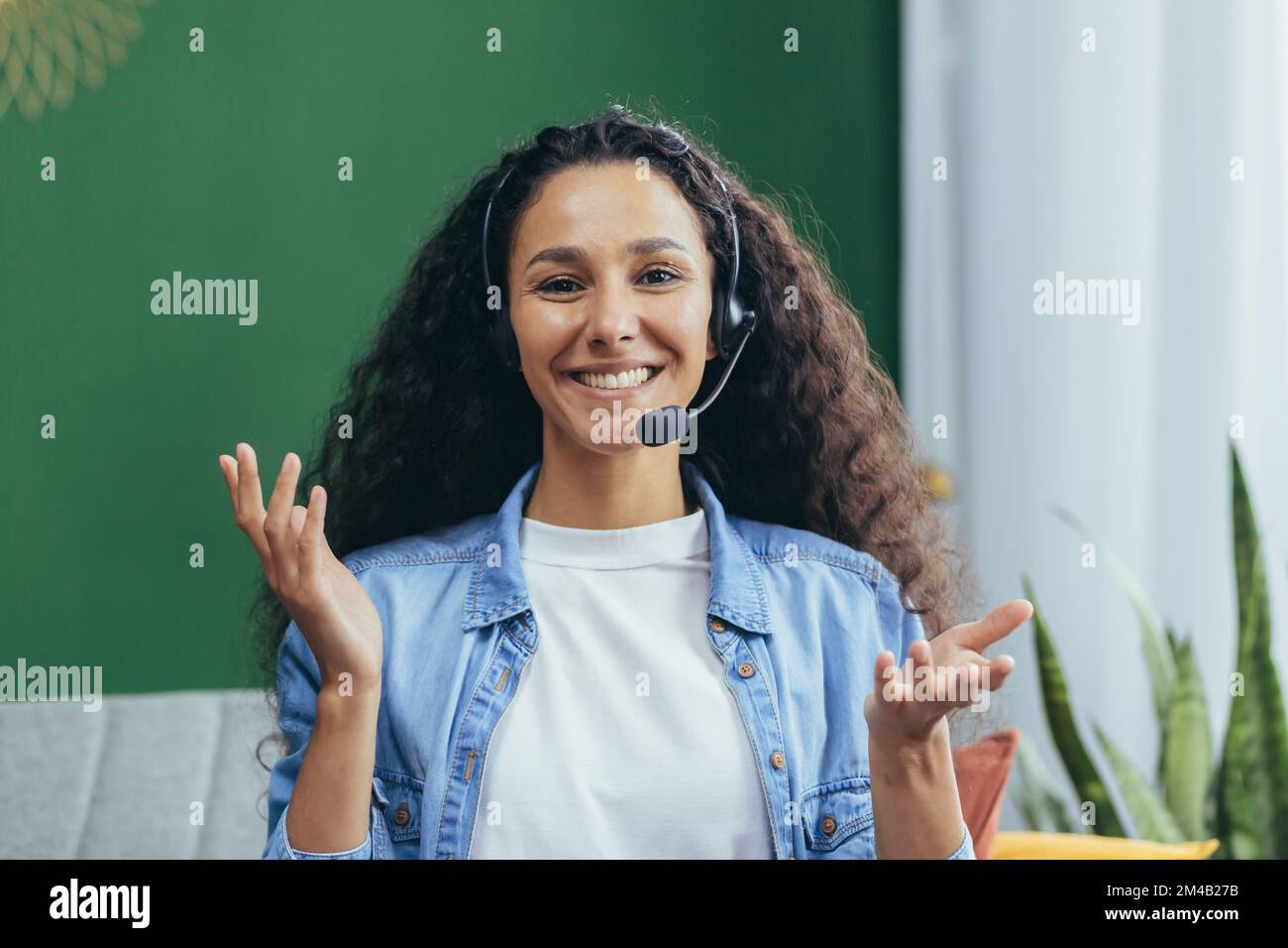 Donna ispanica gioiosa con cuffie per videochiamate a casa sorridente e guardando la telecamera, donna che studia e lavora online seduto a distanza sul divano a casa. Foto Stock