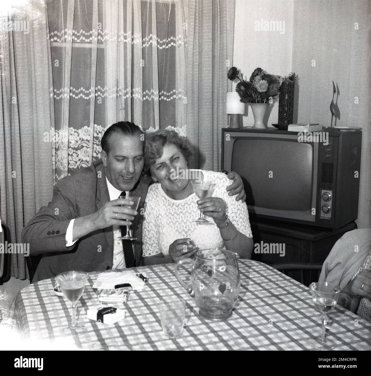 Anni '1960, tempo storico, sera e un uomo maturo e una signora seduti a un tavolo in una sala d'ingresso, un bicchiere di vino in mano, un pacchetto di sigarette sul tavolo, tende a rete sulla finestra e un televisore dell'epoca nell'angolo, Inghilterra, Regno Unito. Foto Stock