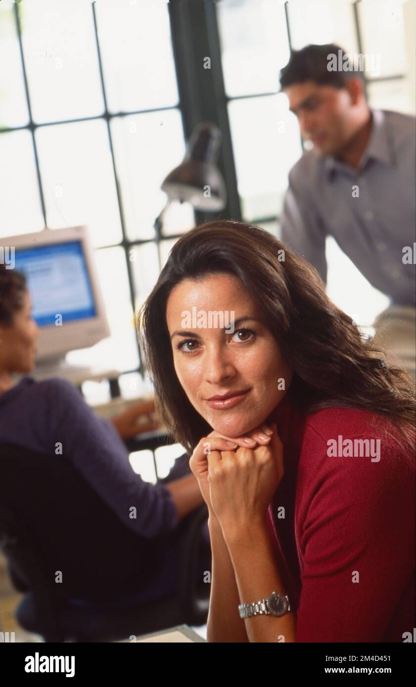 Giovane donna seduta in ufficio con le mani sul mento mentre guarda verso la macchina fotografica con altre persone in ufficio sullo sfondo Foto Stock