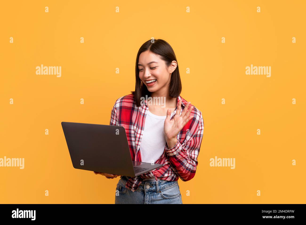 Videochiamata online. Donna asiatica felice che agita per la webcam che tiene e utilizza il computer portatile su sfondo giallo Foto Stock