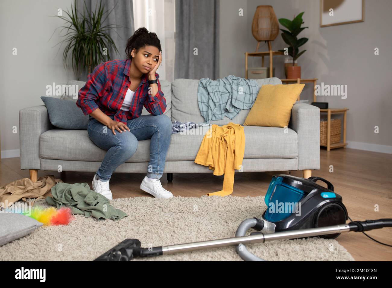 Stanco triste millenario ragazza afroamericana sedersi sul divano, guardare l'aspirapolvere e soffrire di caos e confusione Foto Stock