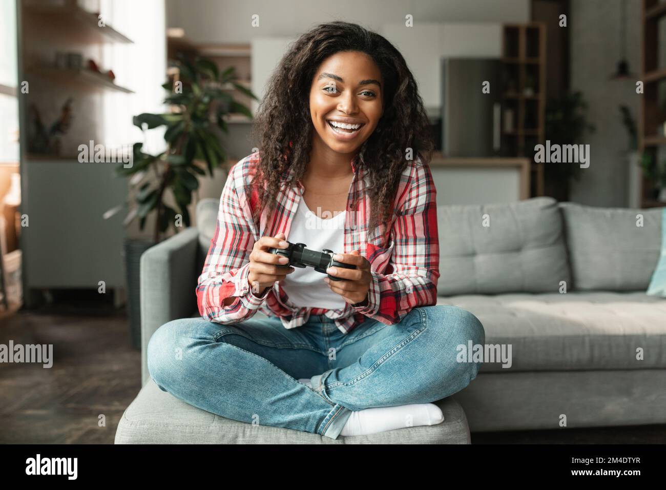 Felice signora nera millenaria con joystick giocare giochi per computer, seduto sul divano da solo, godere di tempo libero Foto Stock