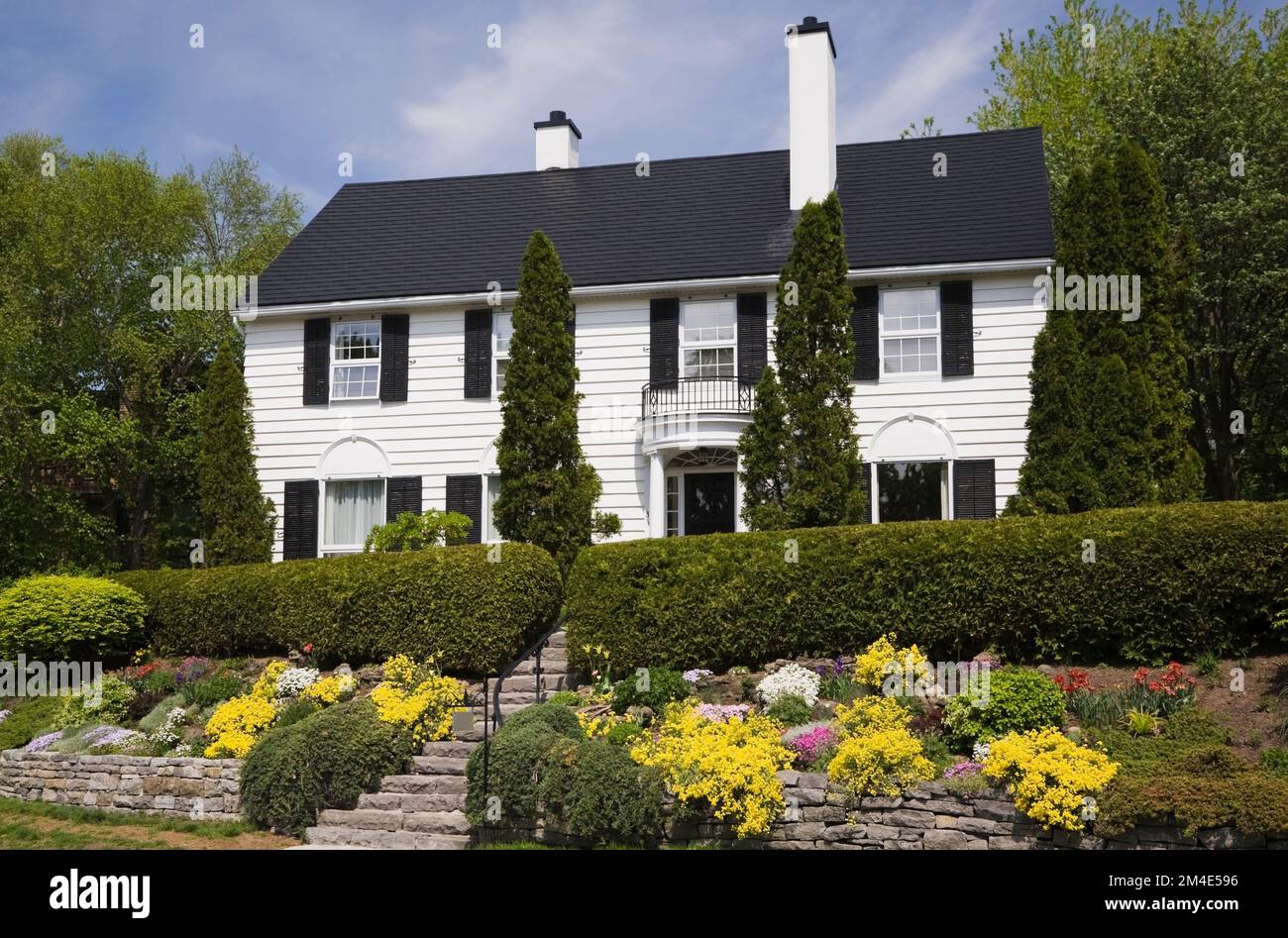 Bordo rialzato con vari arbusti e fiori perenni davanti a un tavolino bianco con finiture nere in stile cottage in primavera. Foto Stock