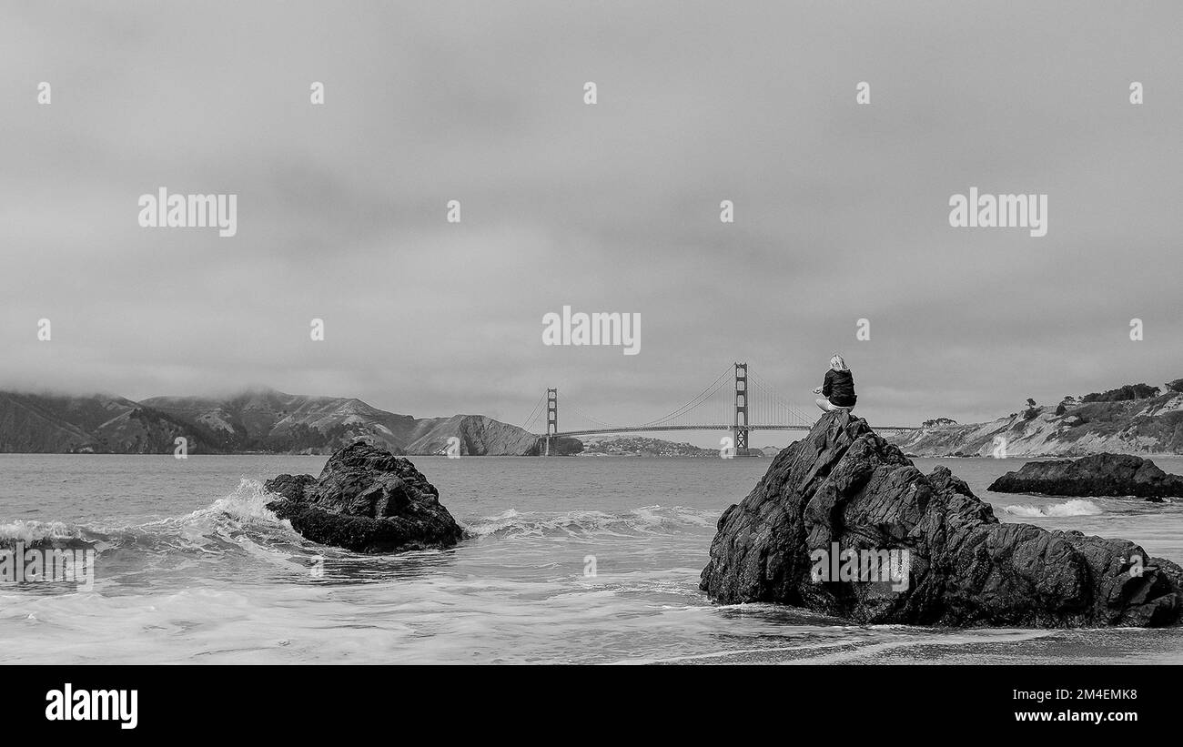 Una donna seduta su una grande roccia sulla spiaggia godendo di una splendida vista del Golden Gate Bridge Foto Stock