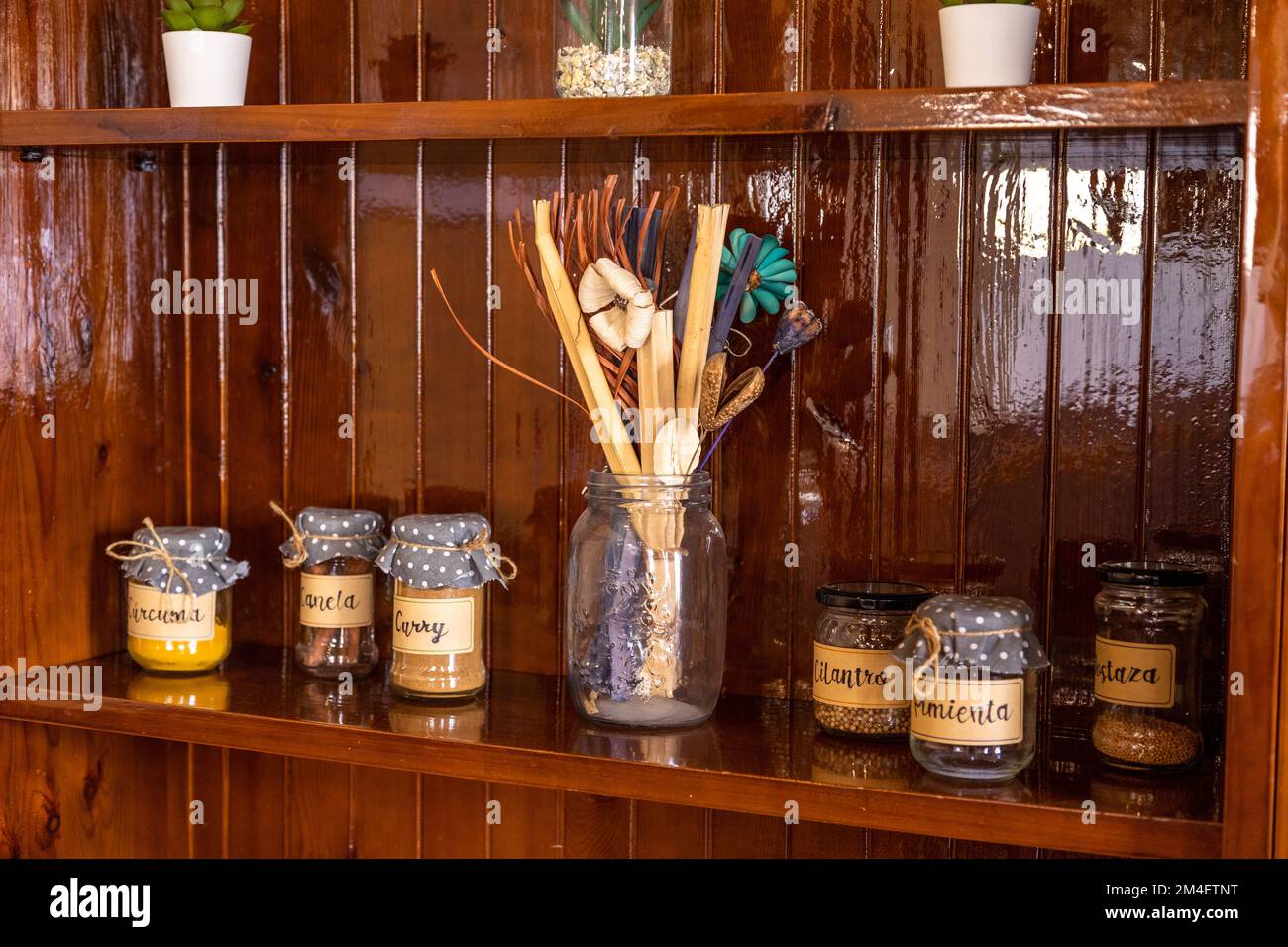 Vasi etichettati con spezie, con panni punteggiati sui coperchi, su una mensola di legno lucido, con fiori artificiali, cucchiai e cannucce in un vaso più grande Foto Stock