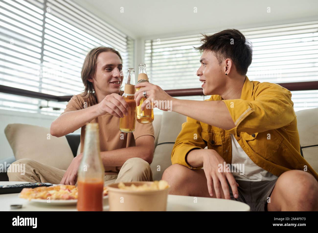 Giovani uomini positivi che si aggraffano le bottiglie quando si beve la birra a casa Foto Stock