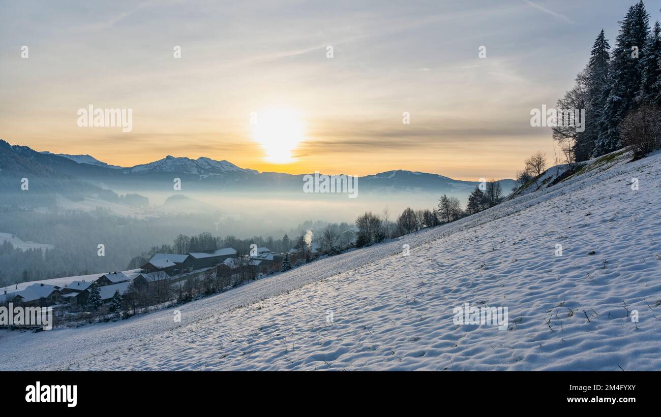 Landschaft, Schnee auf Wiese, Dorf, einzelner Baum und die Berge des Bregenzerwald im Hintergrund. Abendsonne über Hittisau Foto Stock