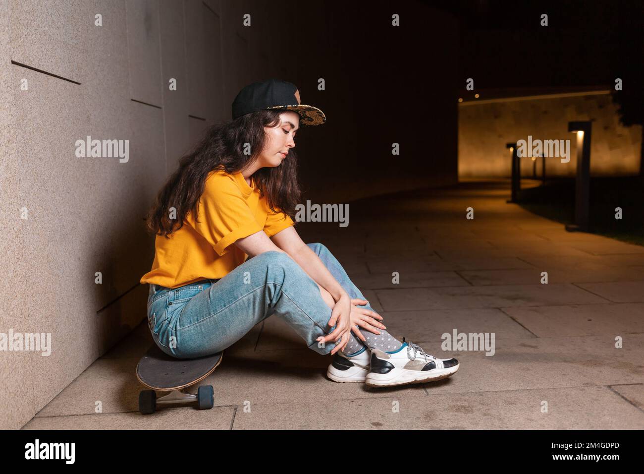 Giovane donna siede su uno skateboard, posando su una strada notturna. All'aperto. Vista laterale. Il concetto di sottocultura giovanile. Foto Stock