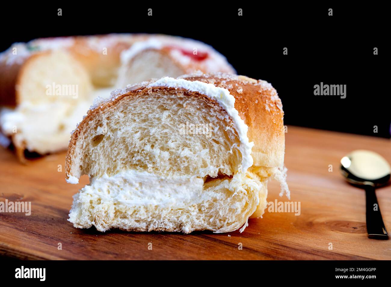 Roscon de reyes farcito con crema, panino o torta tipica della Spagna il giorno dei tre Re con una porzione su un piatto nero e sfondo nero. Foto Stock