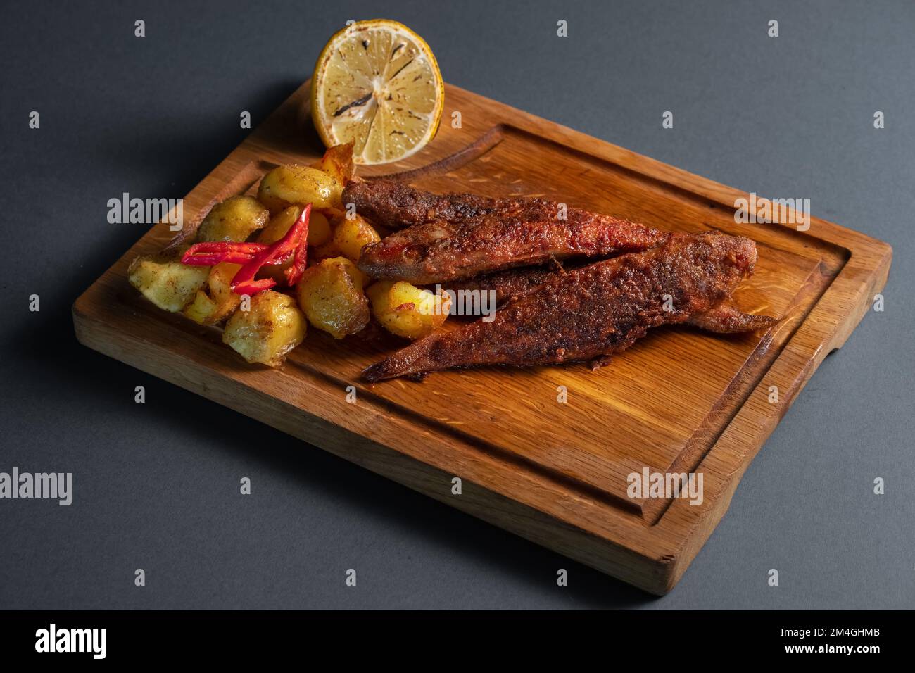 pesce surmullet fritto con patate e limone su tavola di legno su sfondo nero Foto Stock