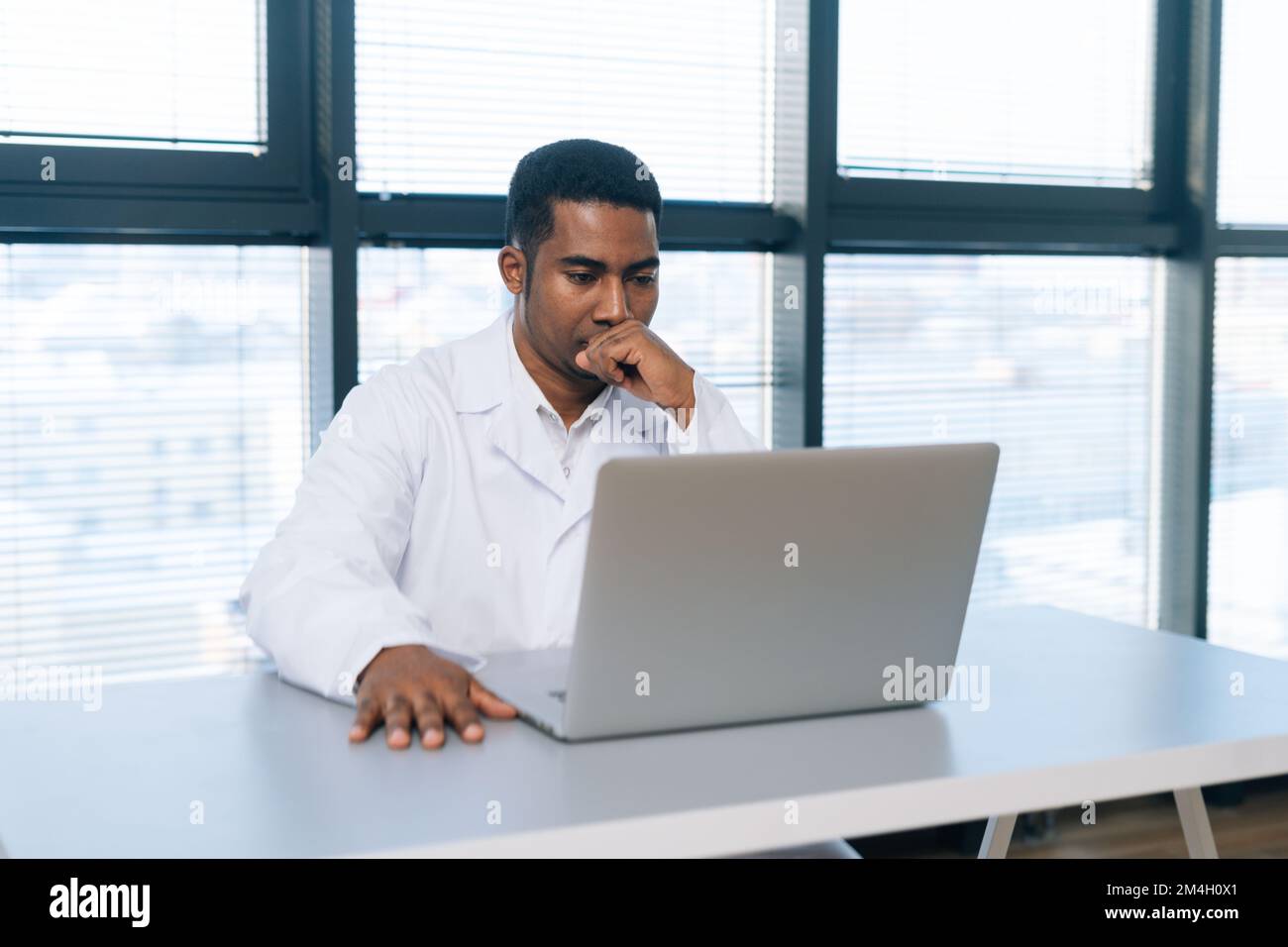 Ritratto del giovane afro-americano medico professionista maschio pensivo in bianco uniforme pensando guardando lo schermo del computer portatile seduto alla scrivania. Foto Stock