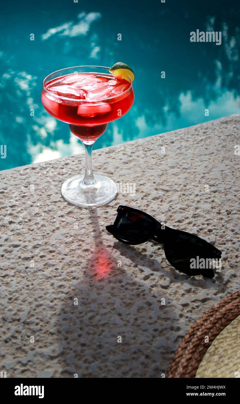 Primo piano di cocktail rosso freddo in piscina con occhiali da sole neri e cappello di paglia Foto Stock
