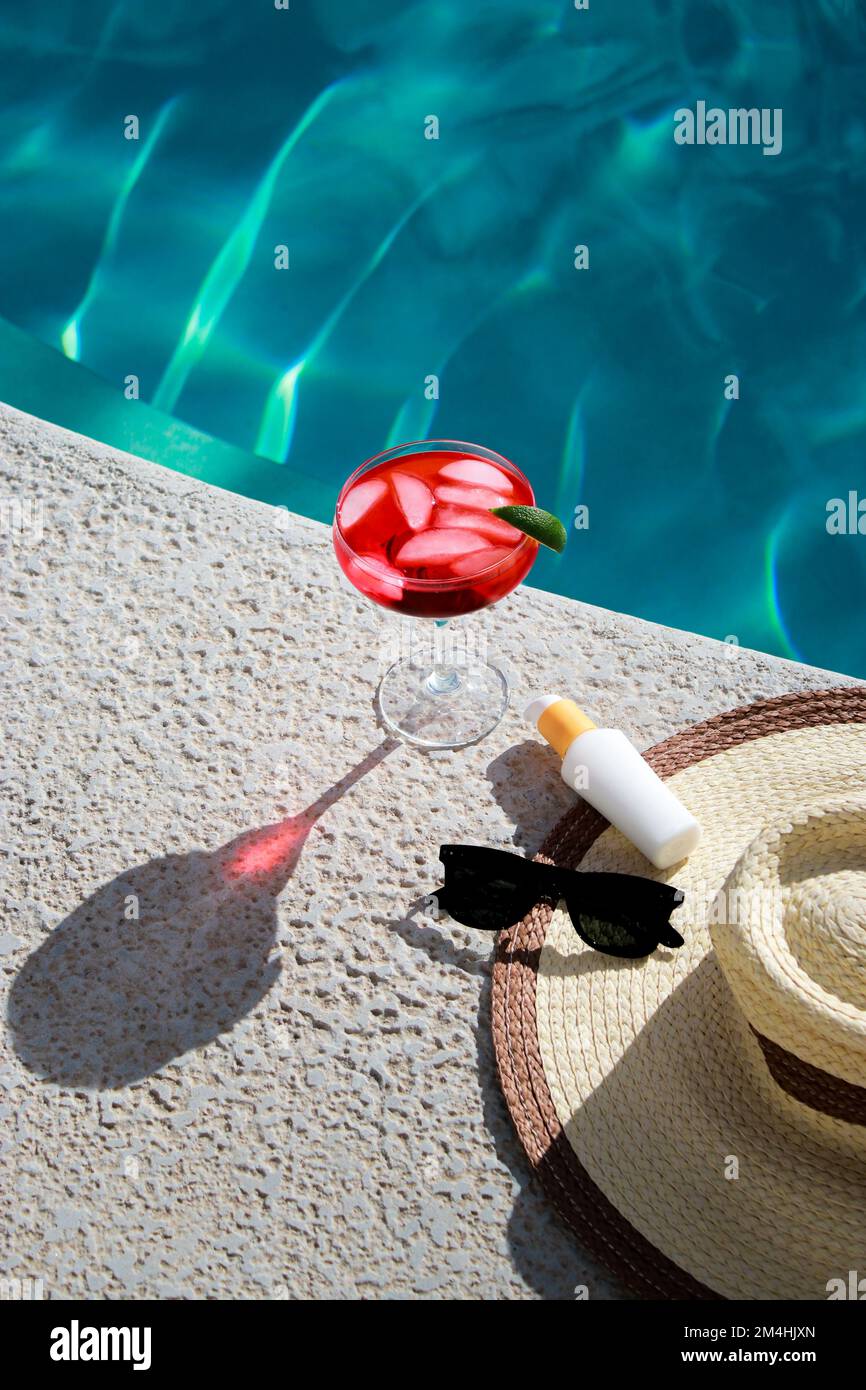 Rinfrescante cocktail rosso in piscina con crema solare, occhiali da sole e cappello di paglia Foto Stock