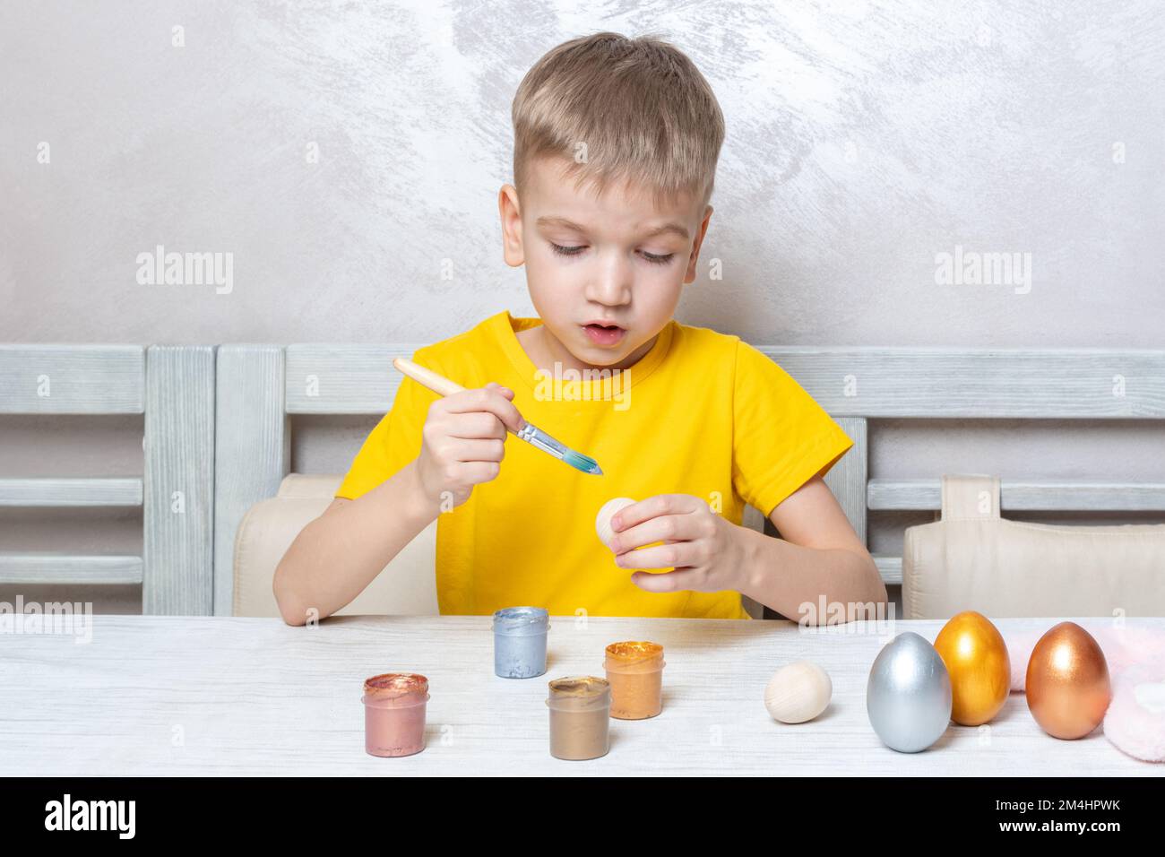 Un ragazzino biondo dipinge le uova per le vacanze di Pasqua a casa. Il bambino si diverte e festeggia la vacanza. Concetto di uova di Pasqua fai da te. Foto Stock