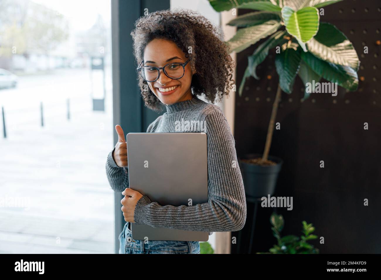 Donna americana sorridente che tiene il laptop e guarda la macchina fotografica mentre si trova vicino alla finestra del caffè Foto Stock