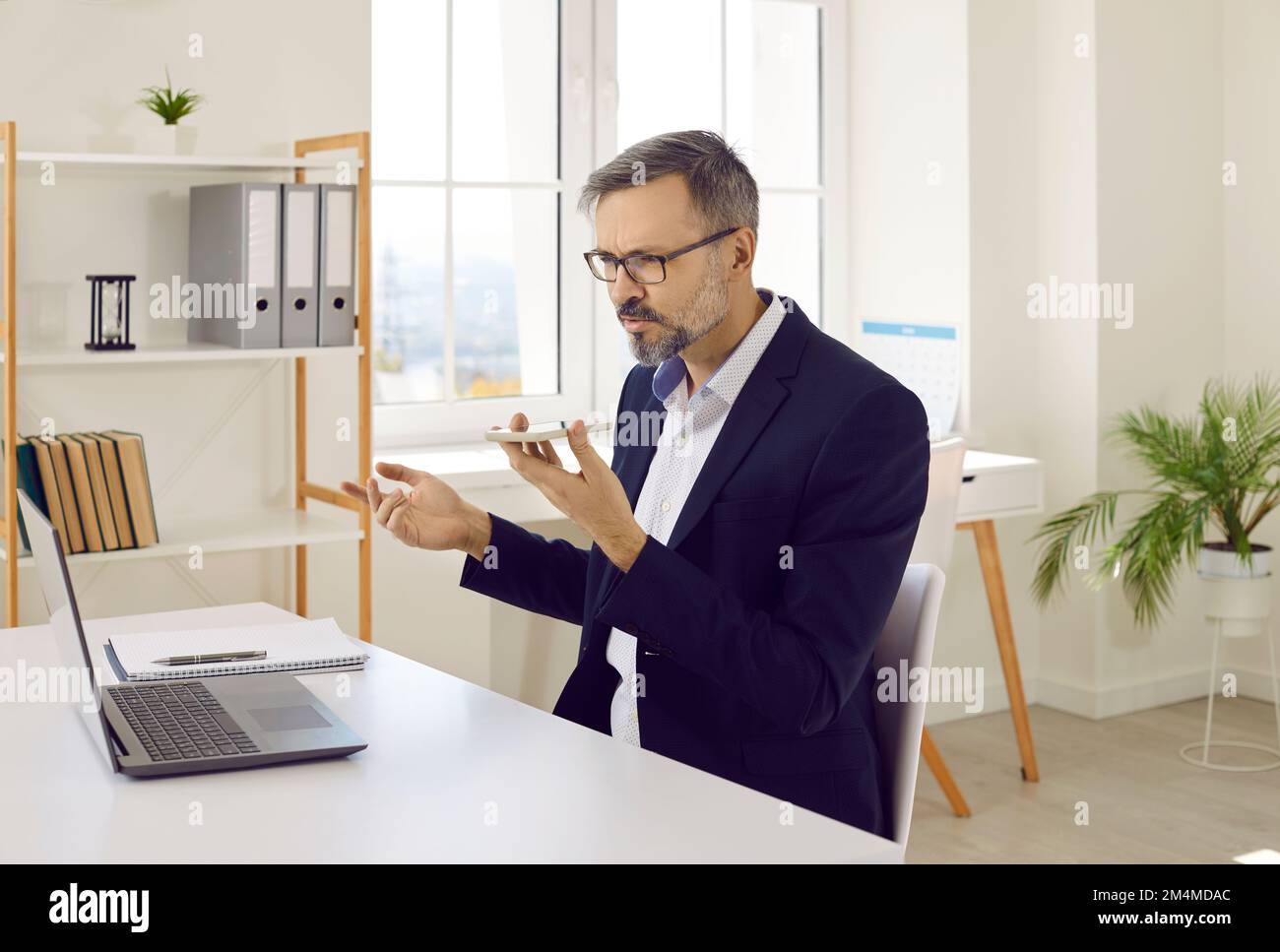 Uomo d'affari che lavora in ufficio, seduto alla scrivania e inviando messaggi vocali sul telefono cellulare Foto Stock