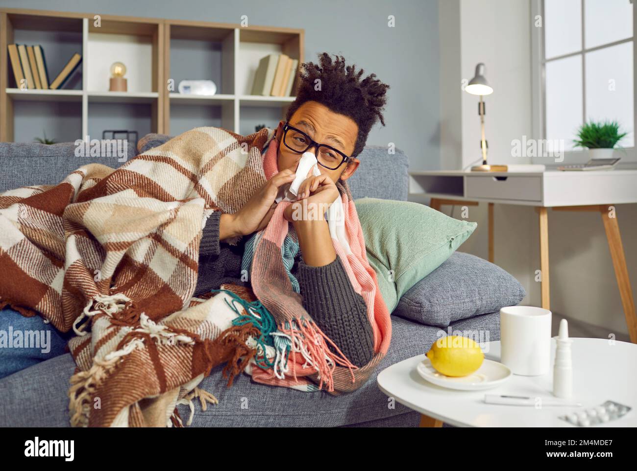 Ammalato afroamericano con un naso che cola sdraiato sul divano e starnuti nel fazzoletto di carta Foto Stock