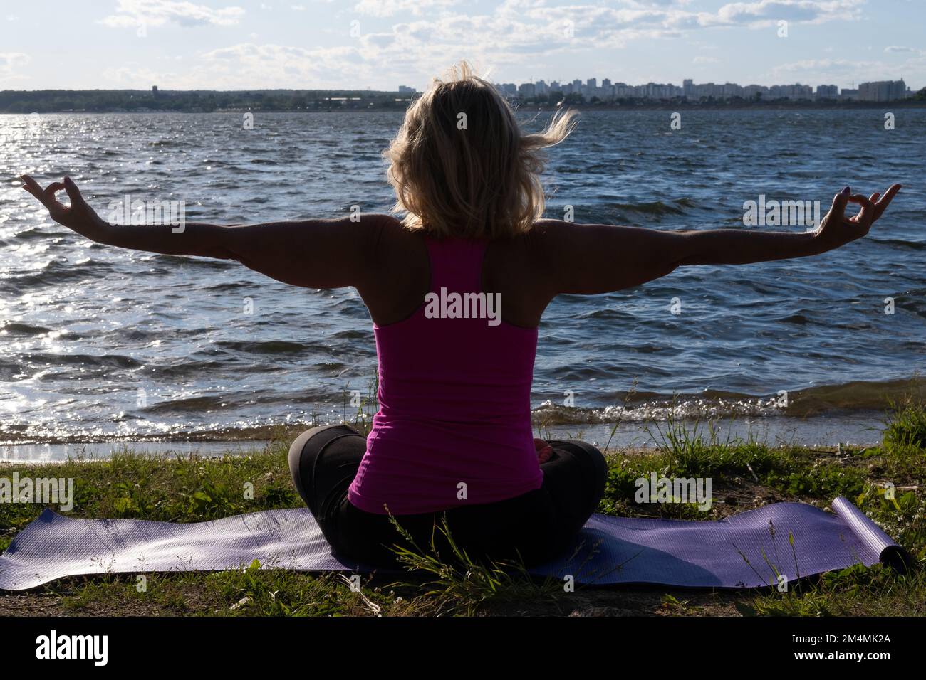 Una donna anziana seduta in posizione loto su un tappetino nel parco e guardando il lago, una vista dal retro. Meditazione. Sport in età avanzata. Yoga Foto Stock