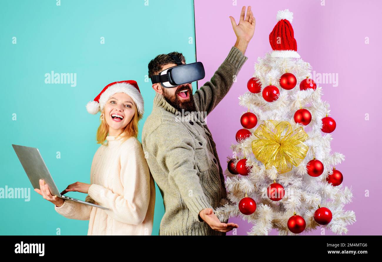 Barbuto uomo in occhiali 3D virtuali e donna in cappello di Santa con notebook vicino albero di natale. Foto Stock