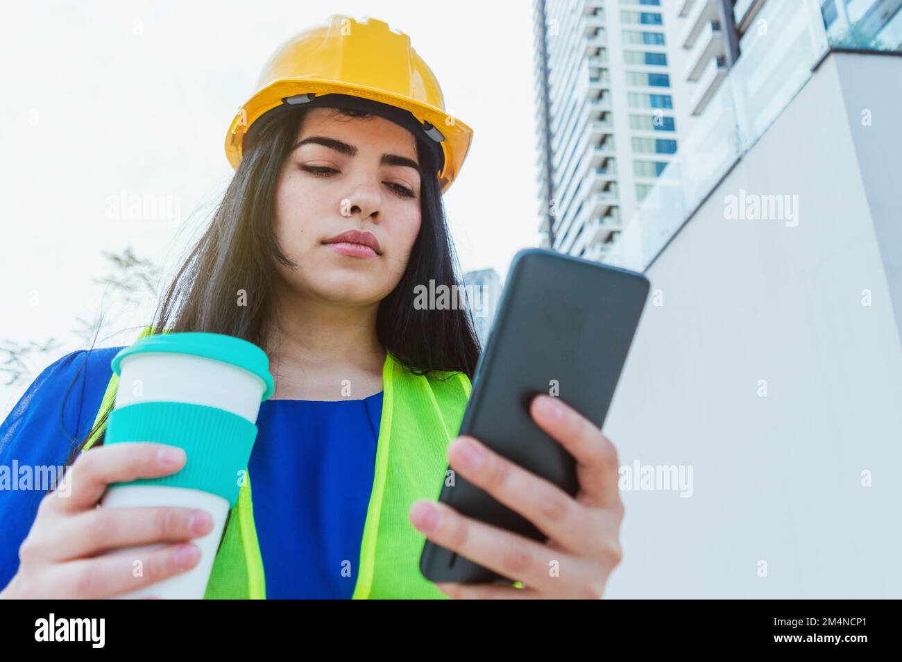 giovane ingegnere caucasico latino donna, vestito in blu con elmetto giallo e giubbotto di sicurezza, in piedi all'aperto bere caffè e utilizzare il telefono durante h Foto Stock