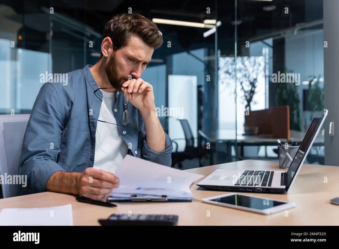 Un giovane serio ragioniere, finanziere, analista, revisore siede nell'ufficio al tavolo. Tiene con cura occhiali, look, analizza account, budget, lavora con documenti e notebook. Foto Stock