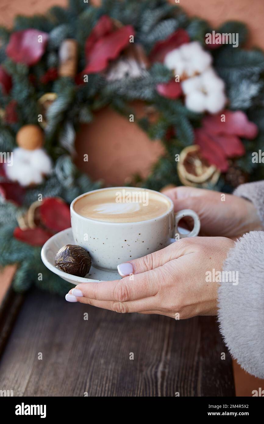 Donna mani tenere una tazza di caffè di fronte alla elegante corona di Natale all'esterno. Accogliente casa di festa. Capodanno mattina. Foto Stock