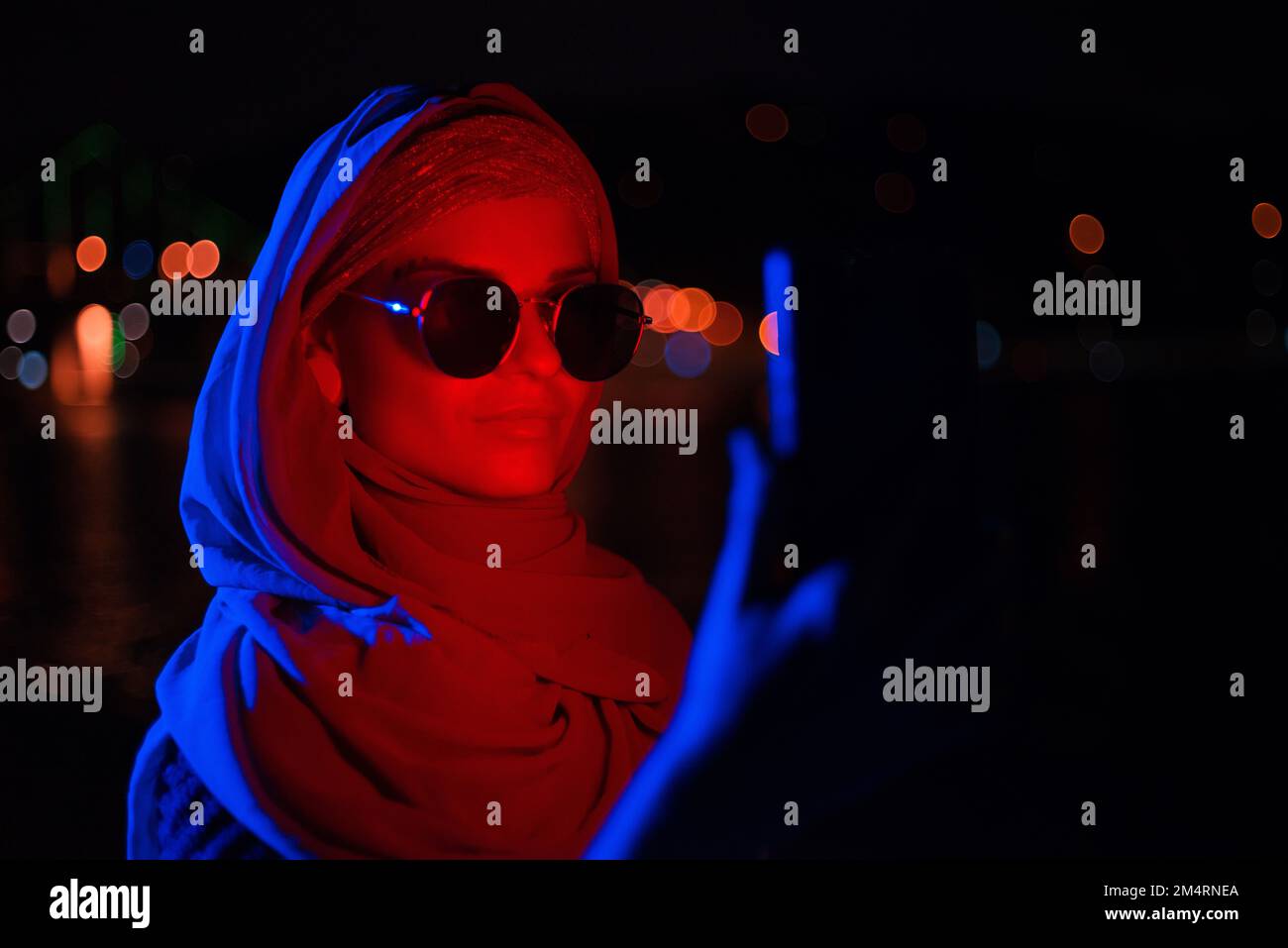 Ritratto di una giovane donna d'affari musulmana che indossa occhiali da sole in Hijab utilizzando uno smartphone moderno con illuminazione al neon rosso illumina il suo volto di notte Foto Stock