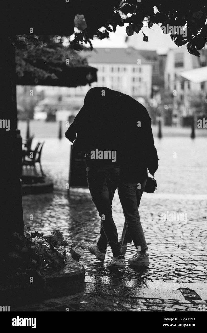 Una scala di grigi verticale di una coppia che cammina mentre scherza sotto una giacca dalla pioggia in Liegi Foto Stock