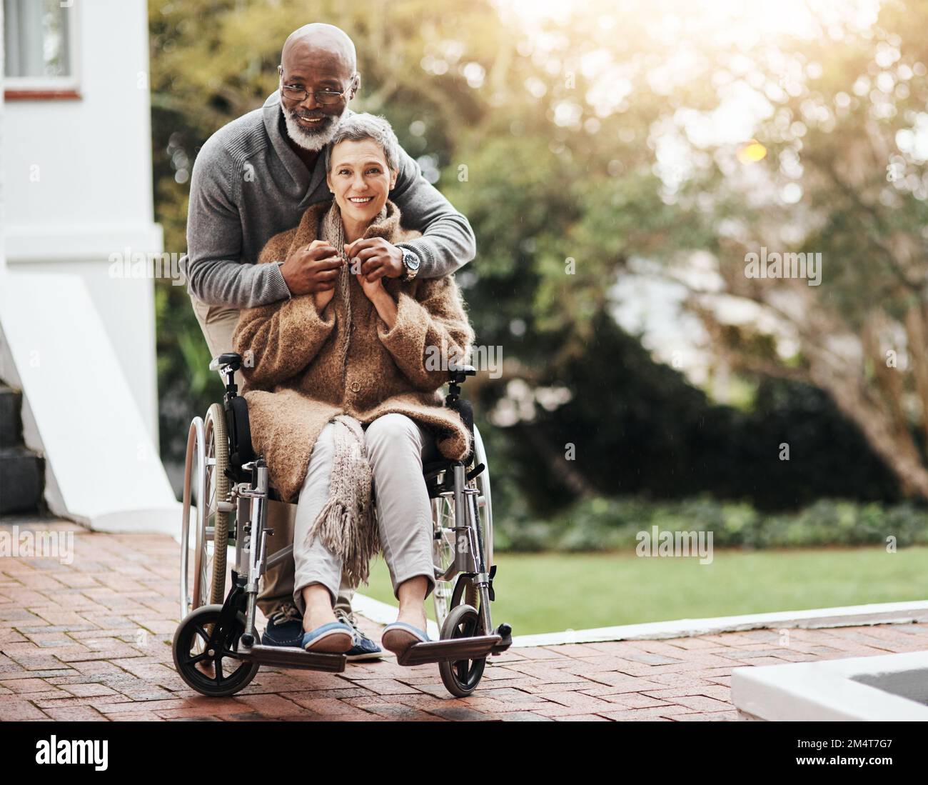 HES è sempre stato al suo fianco attraverso tutto. un uomo anziano che spinge la moglie in sedia a rotelle intorno al cortile a casa. Foto Stock