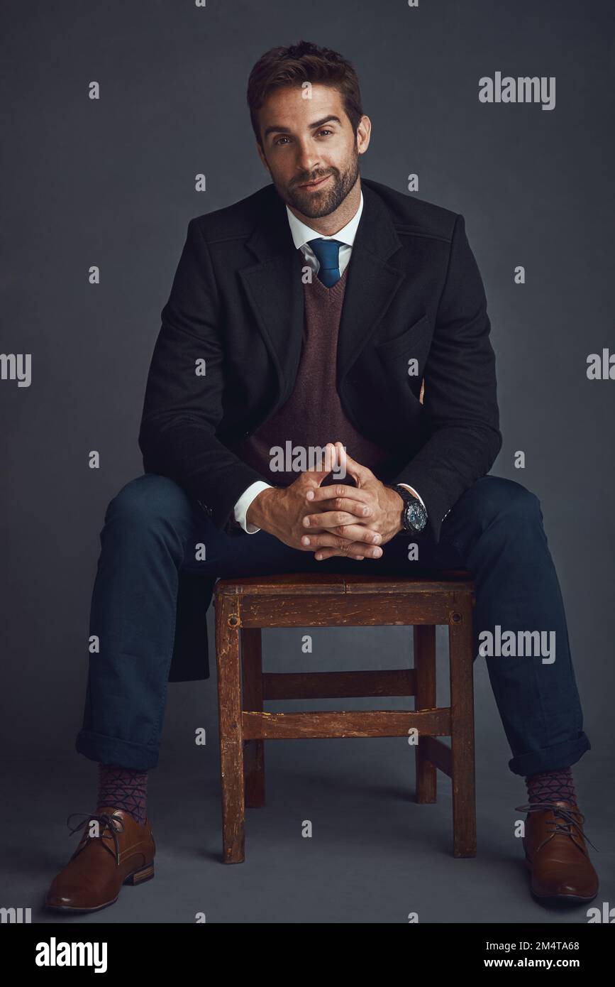 L'esperto di stile. Studio ritratto di un giovane uomo elegantemente vestito seduto su una sedia su uno sfondo grigio. Foto Stock