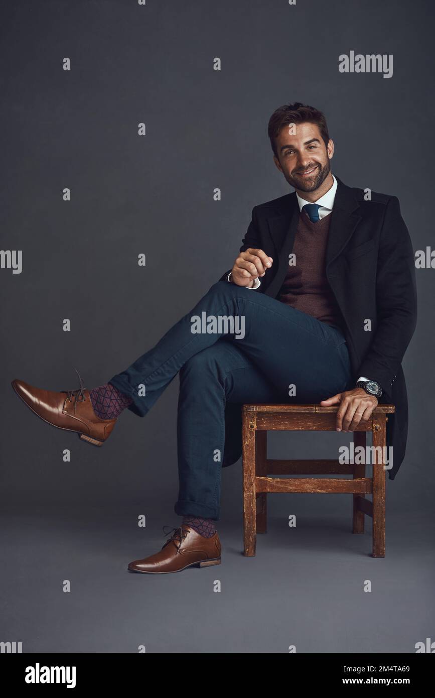 Mi piace mantenere il mio look confortevole ed elegante. Studio ritratto di un giovane uomo elegantemente vestito seduto su una sedia su uno sfondo grigio. Foto Stock