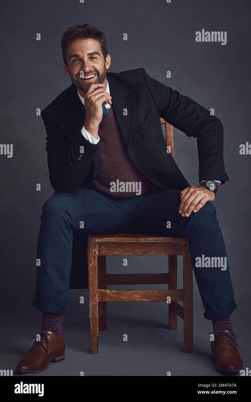 Moda per il lavoro e dopo il lavoro. Studio ritratto di un giovane uomo elegantemente vestito seduto su una sedia su uno sfondo grigio. Foto Stock
