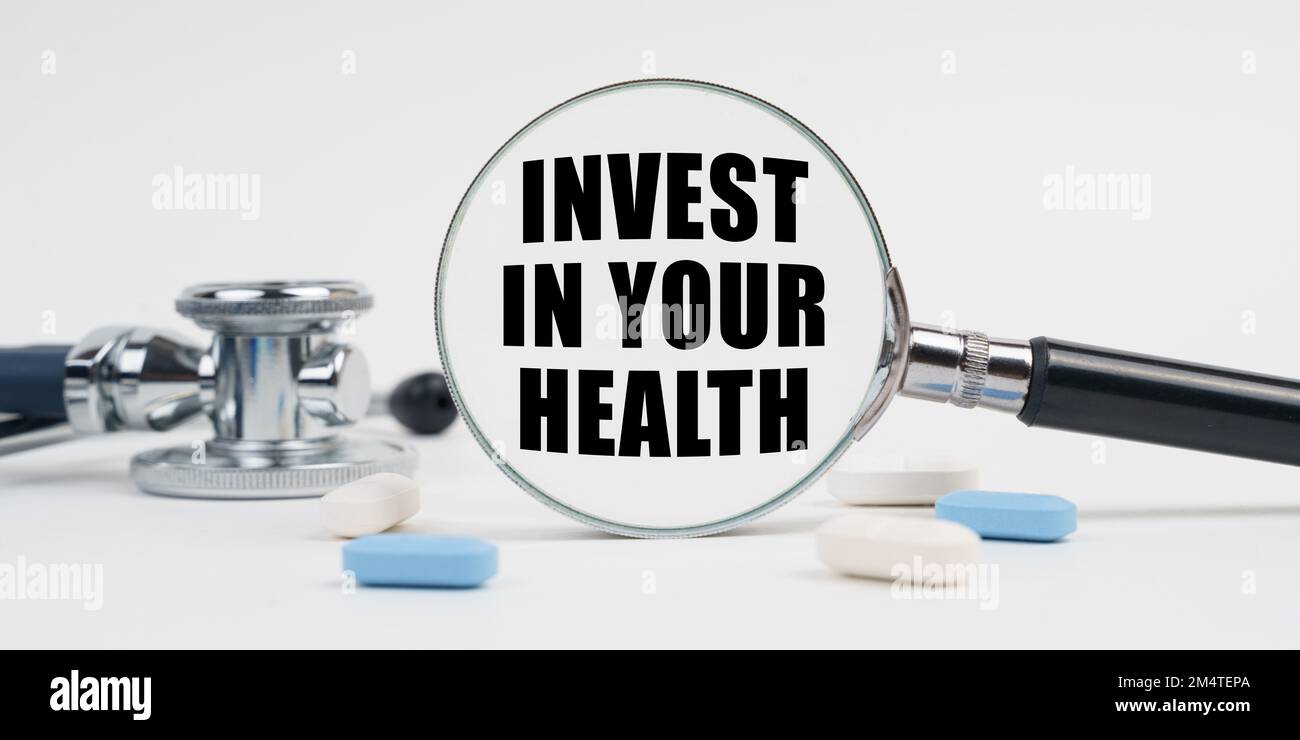Medicina e concetto di salute. Su una superficie bianca si trovano pillole, uno stetoscopio e una lente d'ingrandimento con l'iscrizione - INVESTIRE NELLA VOSTRA SALUTE Foto Stock