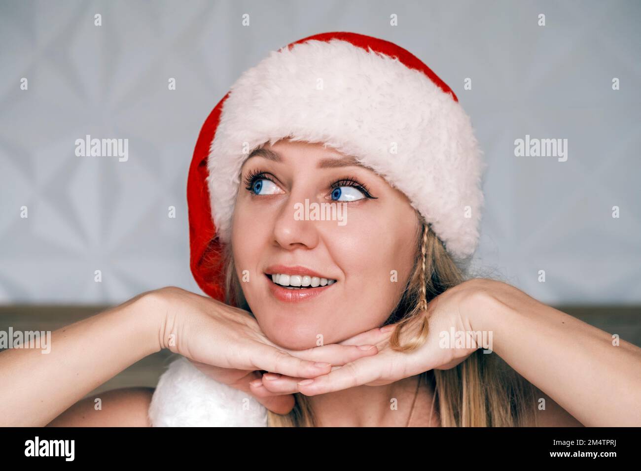 Sorridente Santa ragazza che indossa cappello rosso sta posando il suo mento su entrambe le mani guardando verso l'alto lato. Ritratto di bella giovane bionda con occhio blu nel cappello di Babbo Natale Foto Stock