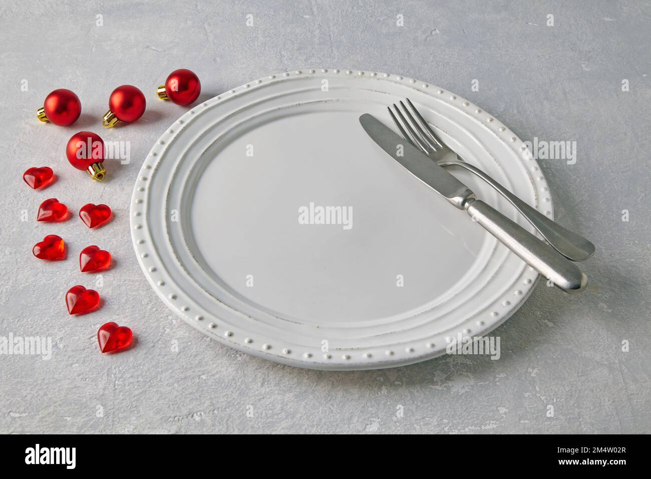 Un piatto bianco vuoto in ceramica con posate circondate da palline rosse di natale e cuori su un tavolo di cemento chiaro Foto Stock