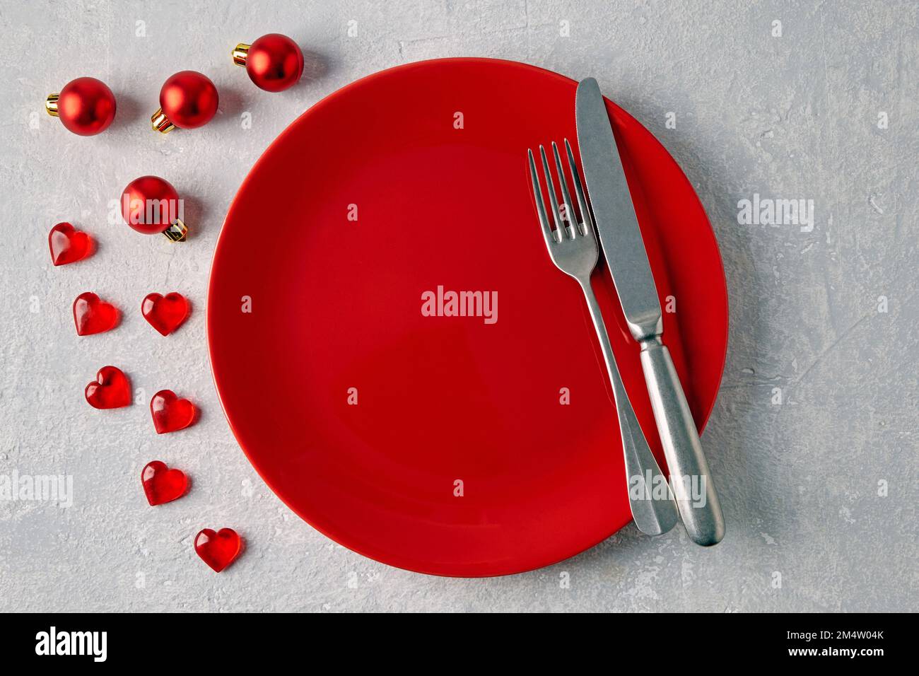 Un piatto di ceramica rosso vuoto con posate circondate da palline rosse di natale e cuori su un tavolo di cemento chiaro Foto Stock